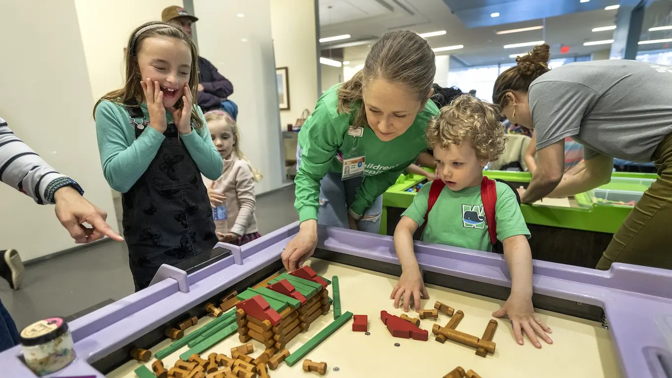 An adult helps young children playing with building blocks in an indoor setting.