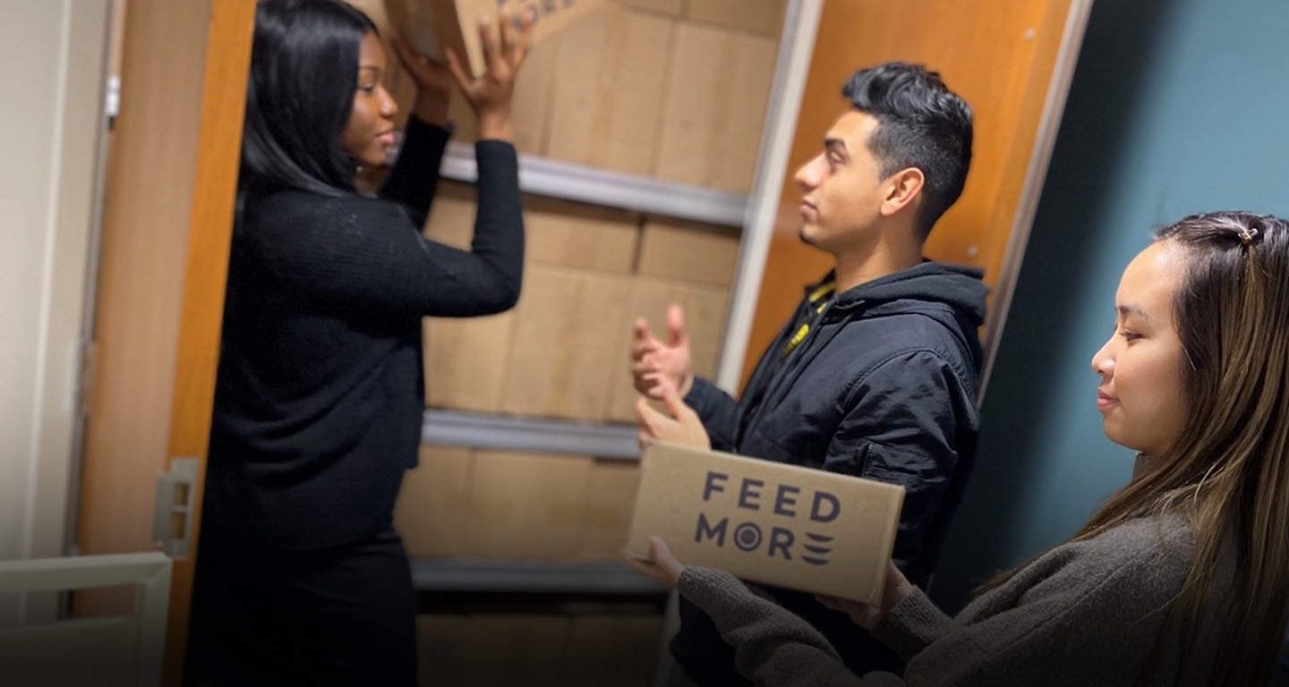Three young people hold Feed More boxes and put them on a shelf.