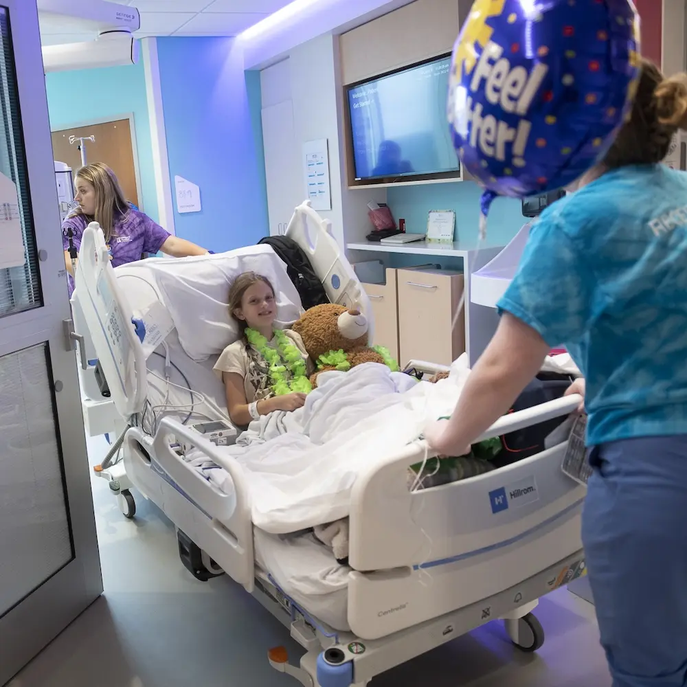 A smiling pre-teen girl and her teddy bear in a hospital bed at the Children’s Tower ar CHoR