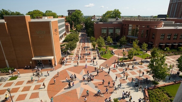 Aerial view of Virginia Commonwealth University’s bustling campus with students walking across the Compass Plaza, surrounded by the red-bricked Hibbs Hall and other campus buildings on a sunny day.