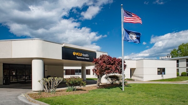 The front entrance of VCU Health Tappahannock Hospital under a bright blue sky. The American and Virginia state flags wave on flagpoles amidst well-kept landscaping.