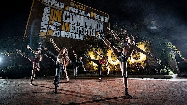 A night scene of dancers performing an expressive routine in front of a vibrant VCU outdoor banner with motivational words.