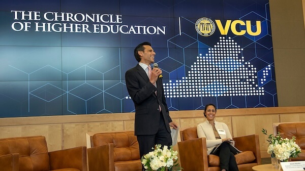 A man standing with a microphone in front of a screen that reads "THE CHRONICLE OF HIGHER EDUCATION," alongside an illustration of Virginia with the letters "VCU" above it. Next to him, a woman is sitting in a brown chair, smiling.