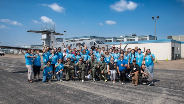 A large group of people in matching blue shirts posing for a group photo in front of a hangar with a research aircraft.