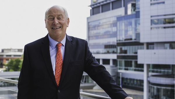 A smiling man in a suit standing on a balcony with modern buildings in the background.