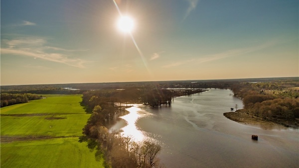 An aerial perspective of the James River adjacent to a field and a row of trees.