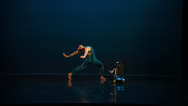 A solo dancer captured mid-performance on a dark stage, , demonstrating a dramatic pose with arms outstretched, and dancing alongside a small robot.
