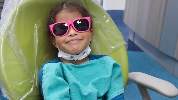 A young girl in a dentist’s chair, wearing sunglasses, and smiling.