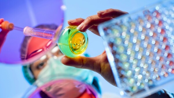 Close-up of a scientist’s hands pouring a yellow liquid into a container, with a multi-well plate and blurred laboratory background.