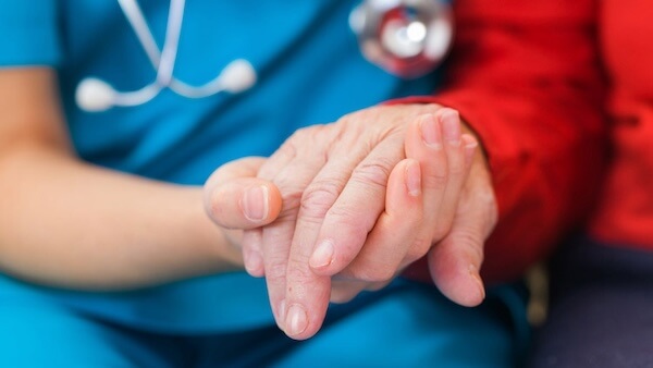 Close-up image of a doctor in blue scrubs holding the hand of an older patient in a red sweater.