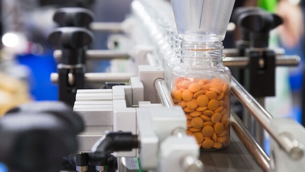 Precision machinery at work in a pharmaceutical manufacturing setting, filling orange capsules into a bottle on a production line.