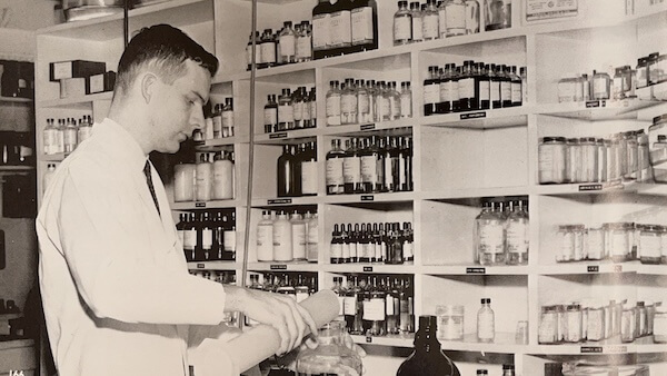 Black and white photo of a man in a lab coat examining a bottle in a vintage pharmacy.