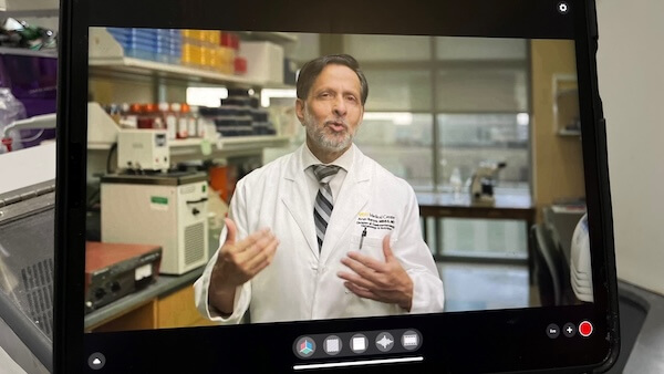 Image captured from a tablet showing Dr. Arun Sanyal, in a white lab coat, speaking and gesturing during a video shoot in a laboratory setting, with equipment and shelves filled with supplies in the background.
